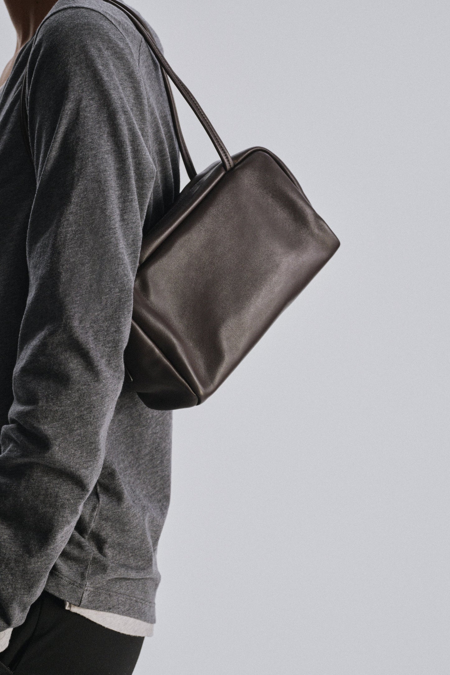 The Row Astra Bag in dark brown worn on the shoulder over a grey long-sleeve top