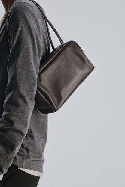 The Row Astra Bag in dark brown worn on the shoulder over a grey long-sleeve top